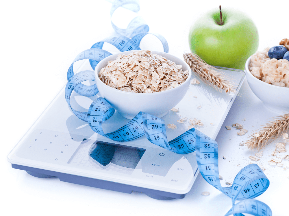 Tigela de aveia, maçã verde e outras frutas sobre uma balança digital, com fita métrica azul enrolada ao redor.